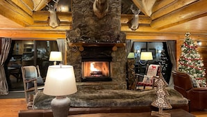 Interior de uma cabana de madeira com lareira de pedra, duas luminárias, um pinheiro de Natal e chifres de veado na parede.
