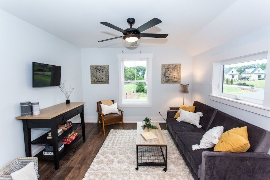 A modern living room with a dark wooden coffee table, a black sofa with yellow cushions, a wooden side table, a black floor lamp, and a ceiling fan.
