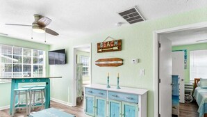 A room with a kitchenette, a dining area with stools, a bed with a view, and a wall with a 'BEACH' sign.