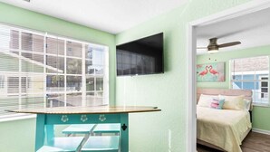 A room with a bed, a TV, a table with chairs, and a window with blinds.
