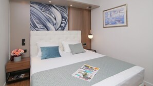 Ein modernes Hotelzimmer mit einem großen Bett, einem Kopfende mit vertikalen Lamellen, einem Wandbild, einem Nachttisch mit Lampe sowie einem gerahmten Bild an der Wand.