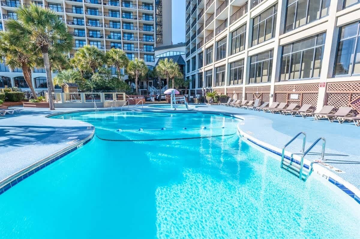 A swimming pool with lounge chairs and palm trees, surrounded by a hotel building with multiple floors and balconies.