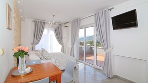 A hotel room with a bed, a TV mounted on the wall, a dining table with chairs, and a view of the outdoors through large windows.