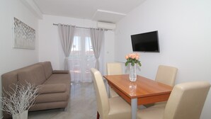 Una sala de estar moderna con un sofá beige, una mesa de comedor de madera con cuatro sillas, un jarrón con flores, un televisor de pantalla plana montado en la pared y una ventana con cortinas de gasa.