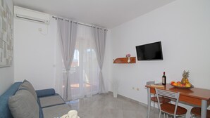 A living room with a sofa, a dining table with chairs, a TV mounted on the wall, and a window with sheer curtains.