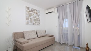 A beige sofa with two pillows, a white wall with a geometric artwork, a white air conditioning unit, sheer curtains, and a small statue on the floor.