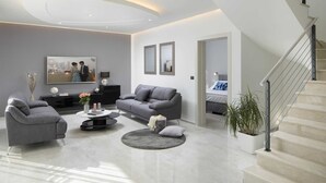 Una sala moderna con un sofá gris, una mesa de centro y un televisor. Se ve una escalera a la derecha y una puerta que da a otra habitación.