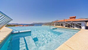Ein klares, blaues Schwimmbecken mit einer gefliesten Terrasse, ein Gebäude mit rotem Ziegeldach und Blick auf ferne Berge.
