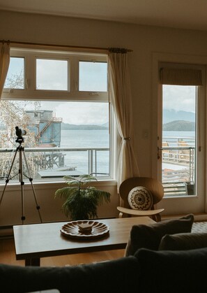 Un salon doté d’une grande fenêtre donnant sur un plan d’eau et un quai. On y trouve un trépied sur lequel est posé un appareil photo, une plante en pot et une table ronde sur laquelle est disposée une assiette décorative.