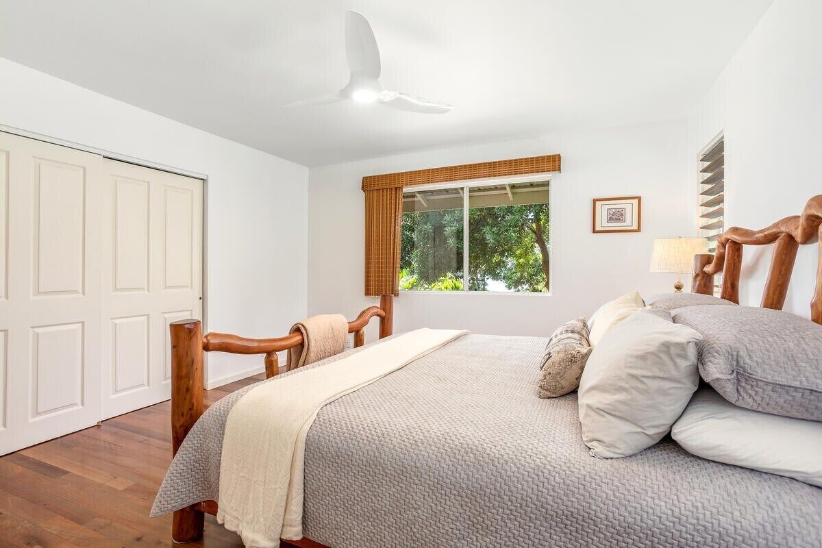 A bedroom with a bed, wooden headboard, a window with a view of greenery, a ceiling fan, and a door.