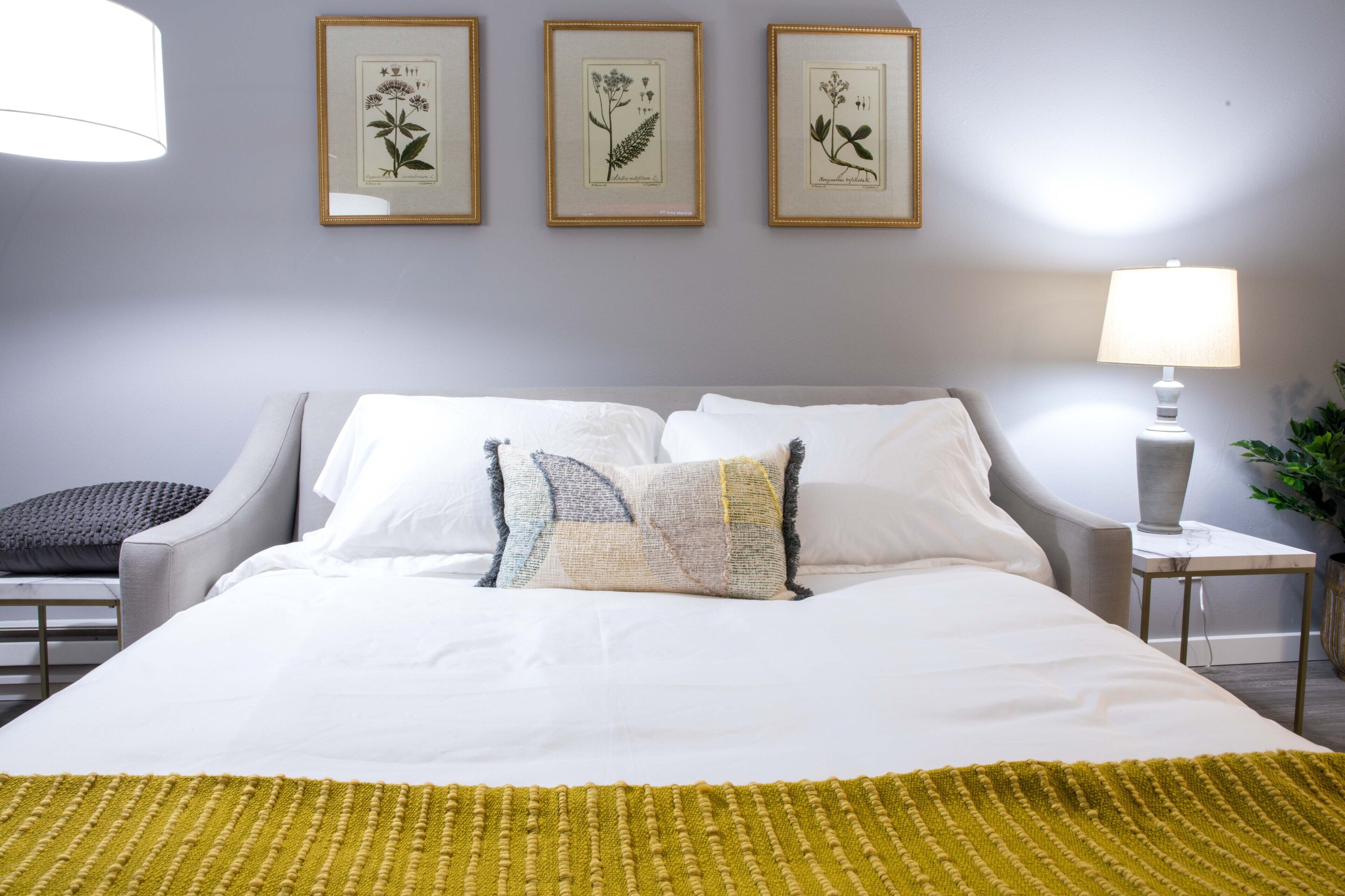 En pænt redt seng med hvide sengetøj og et gult plaide, en grå polstret stol og et natbord med en lampe.