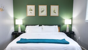 Ein ordentlich bezogenes Bett mit weißen Bettwäsche und einer blauen Zudecke, begleitet von zwei Nachttischen mit Lampen, an einer grünen Wand mit gerahmten Pflanzenbildern.