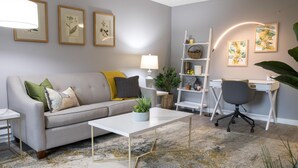 Ein Wohnzimmer mit einer Couch, Couchtisch, Schreibtisch und Stuhl. An den Wänden befinden sich gerahmte Pflanzenbilder und auf dem Schreibtisch steht eine Lampe.