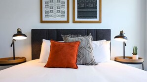 Una cama bien hecha con ropa de cama blanca, dos almohadas y un plaid de punto gris. Hay dos mesitas de noche con lámparas y una pequeña planta en maceta.