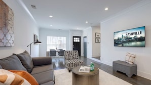 Una sala de estar moderna con un sofá gris, un sillón estampado y una mesa redonda de centro con una revista. Un televisor de pantalla plana muestra “Welcome to Nashville” y un paisaje urbano.