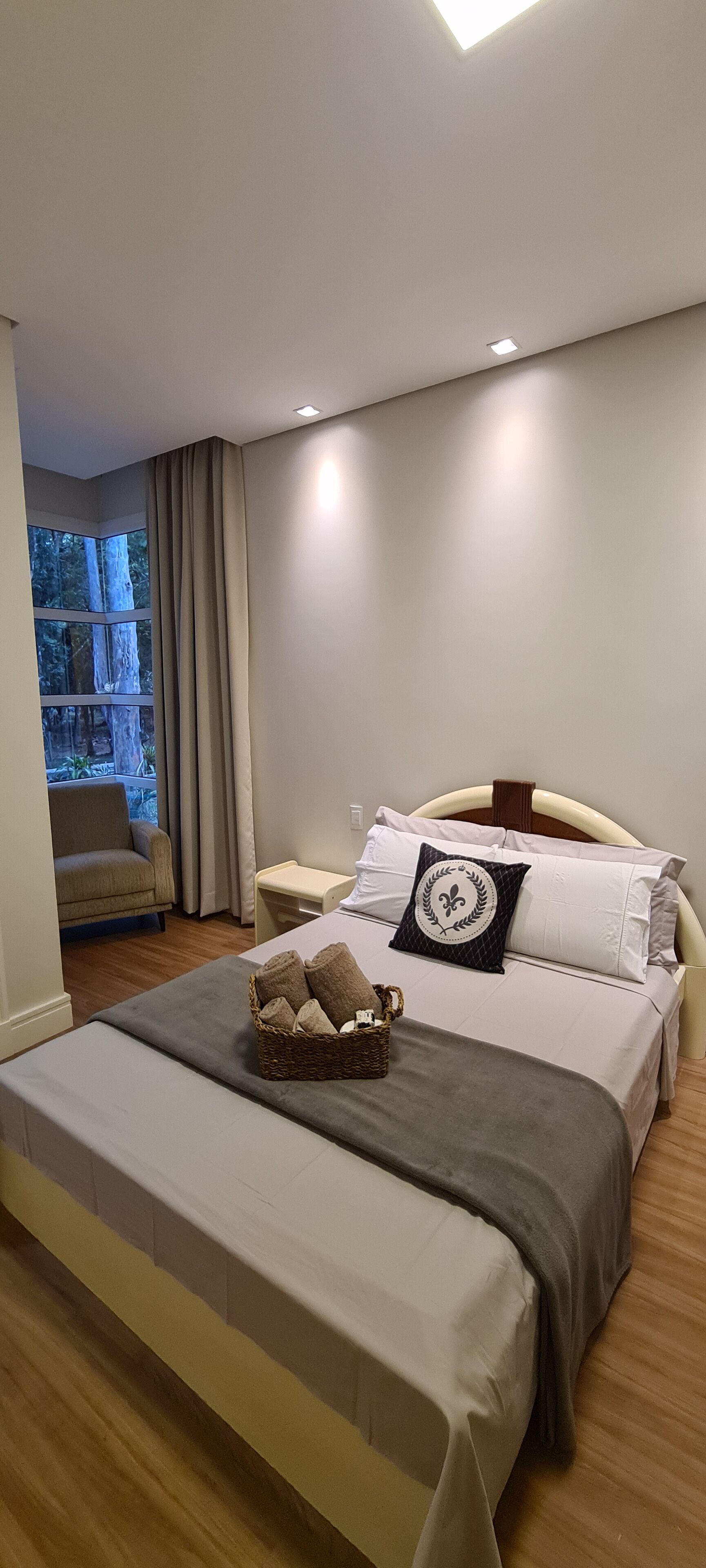 Une chambre à coucher avec un grand lit, une table de chevet, une commode, une fenêtre avec des rideaux et une vue sur les arbres à l’extérieur.