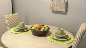 Een eettafel met een fruitschaal, twee groene bekers en een decoratieve wandversiering.