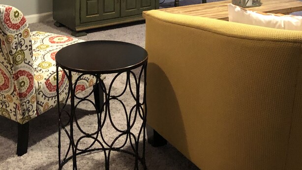 Een woonkamer met een televisie op een kast, een gestreept fauteuil, een gele bank en een kleine ronde tafel.