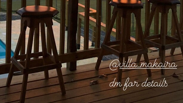 A wooden deck with two stools, overlooking a sunset over a body of water with palm trees.