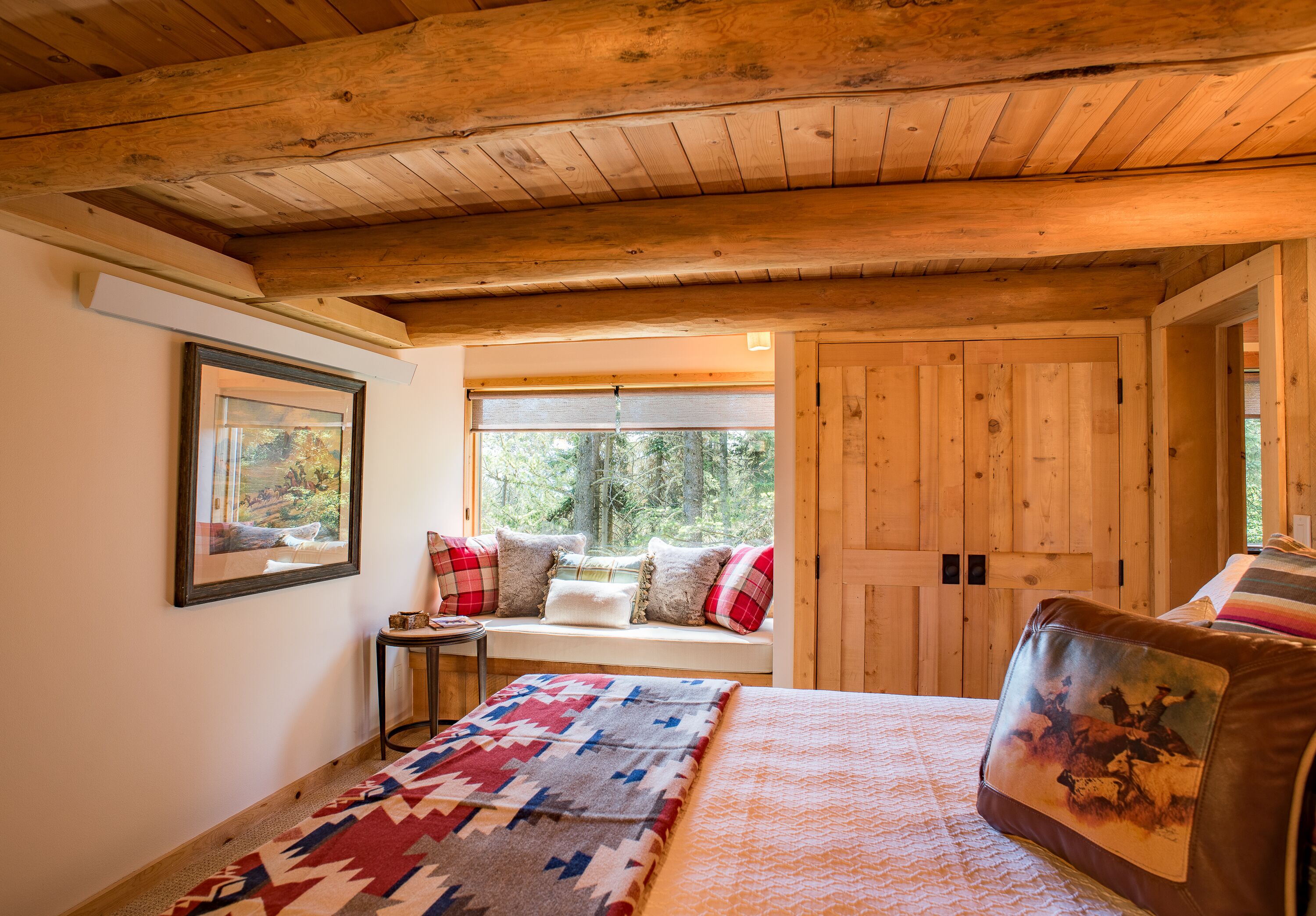 A cozy room with a wooden ceiling, a large bed with a patterned blanket, a window seat with pillows, and a framed picture on the wall.