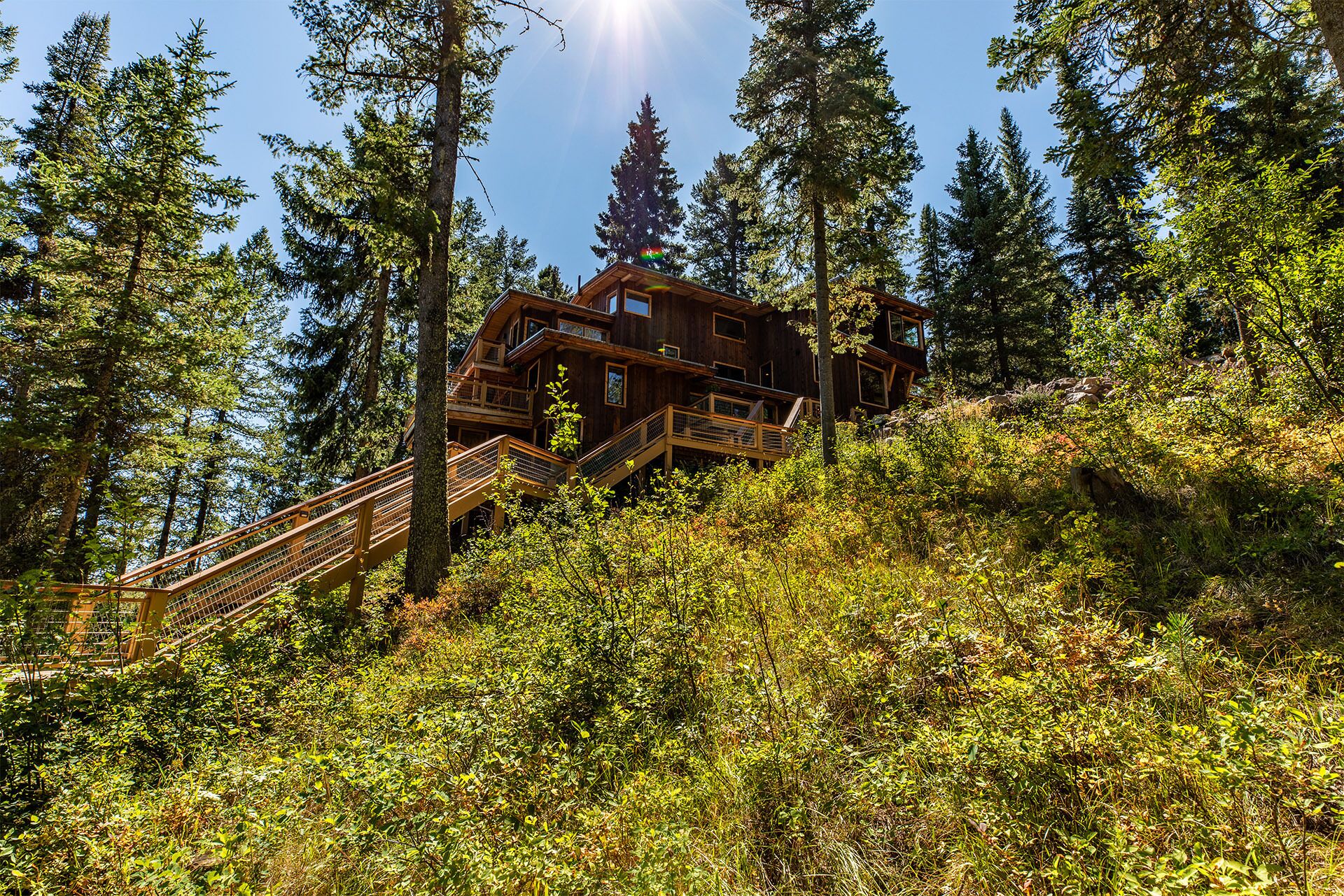 A wooden cabin with multiple balconies, surrounded by dense forest and a wooden staircase leading up to it.