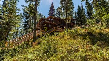 A wooden cabin with multiple balconies, surrounded by dense forest and a wooden staircase leading up to it.