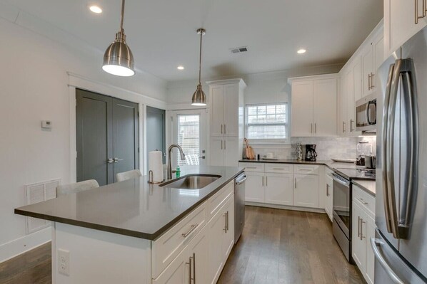 A modern kitchen with white cabinets, stainless steel appliances, a large island with a sink, pendant lighting, and a window providing natural light.