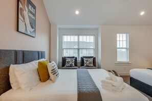Quarto de hotel com uma cama grande, um sofá, duas janelas com persianas e uma luminária no teto.