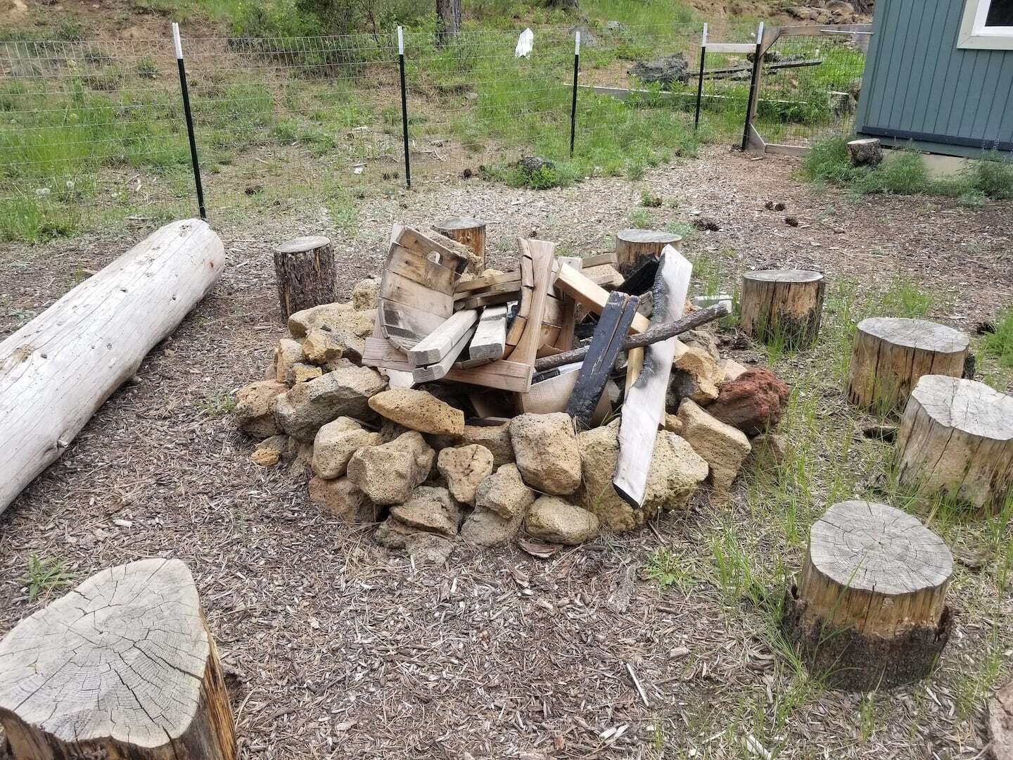Een vuurplaats omringd door rotsen en houtblokken, met een houten paal en een metalen hek op de achtergrond.