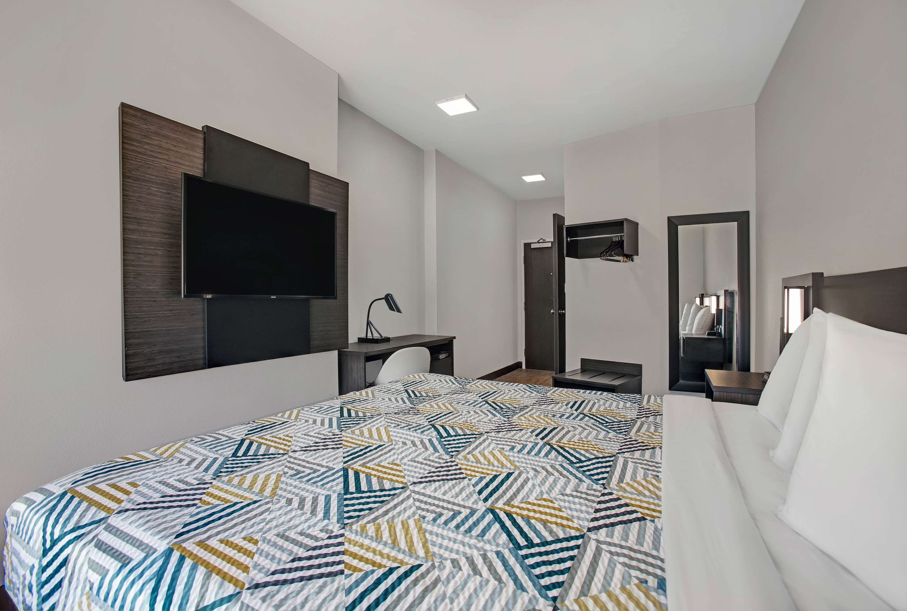 A modern hotel room with a large bed, a flat-screen TV mounted on the wall, a desk with a chair, a mirror, and a wardrobe.