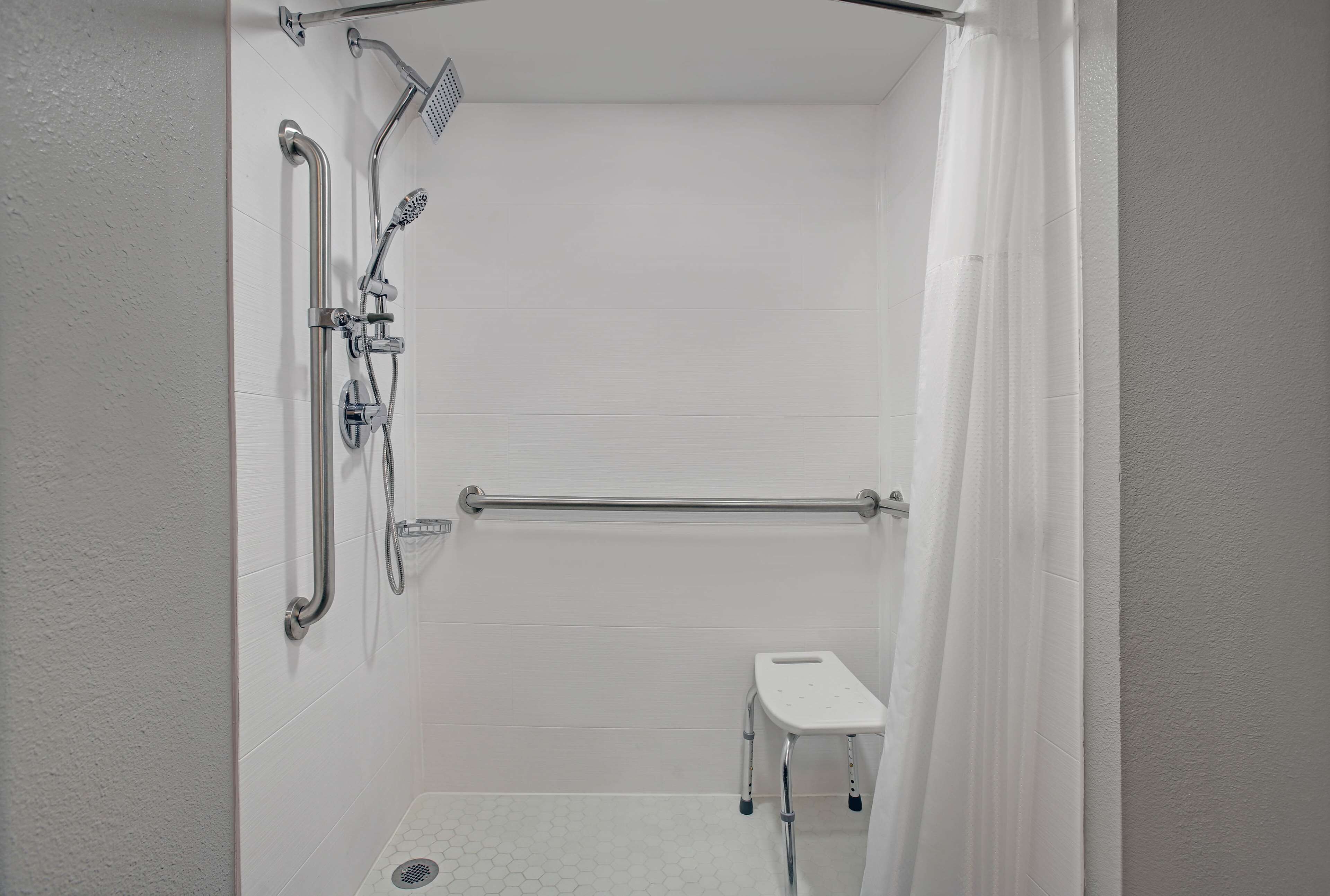 A small, white, tiled shower room with a showerhead, grab bar, and a step stool.