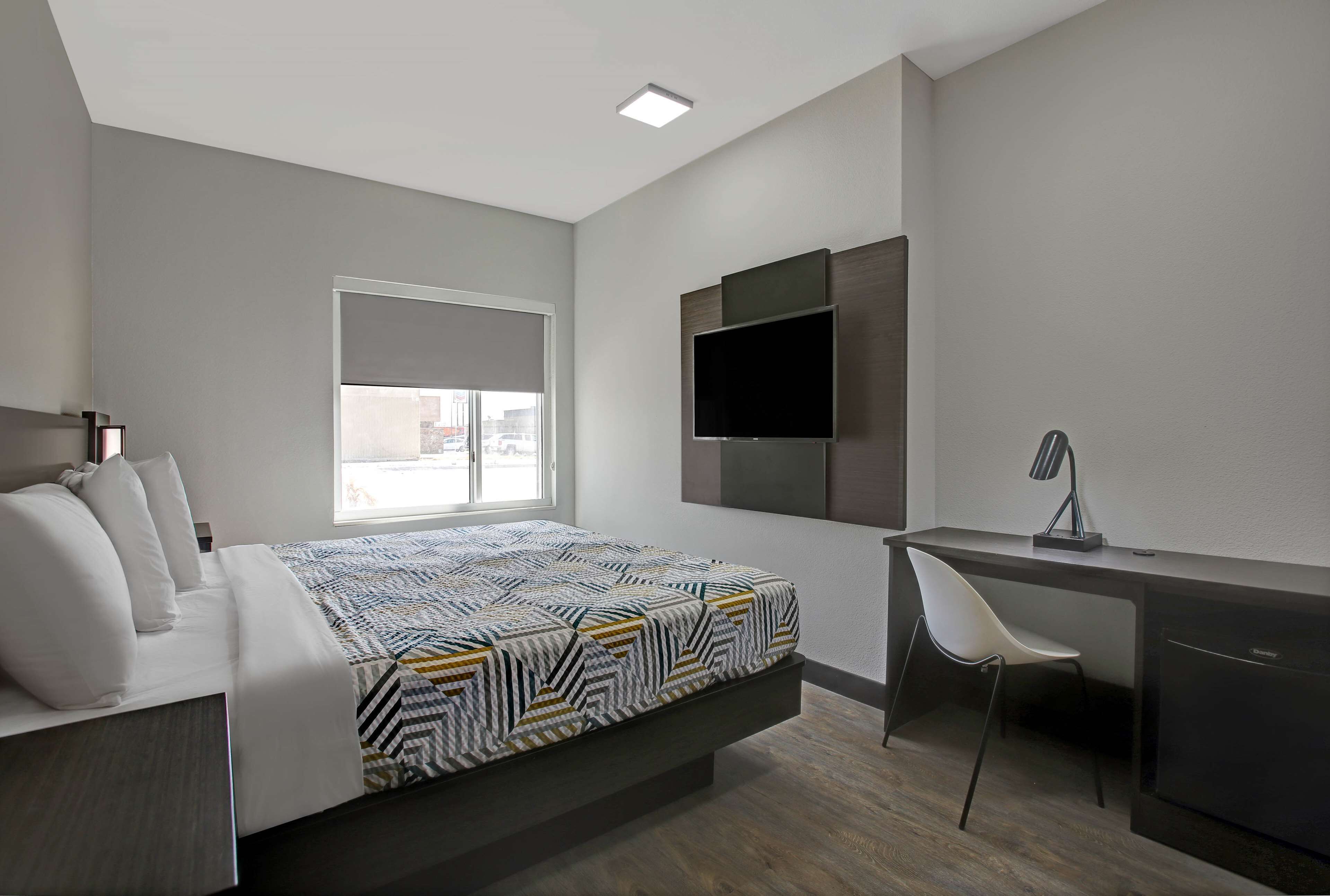 A modern hotel room with a bed, a desk, a chair, a TV, and a window.