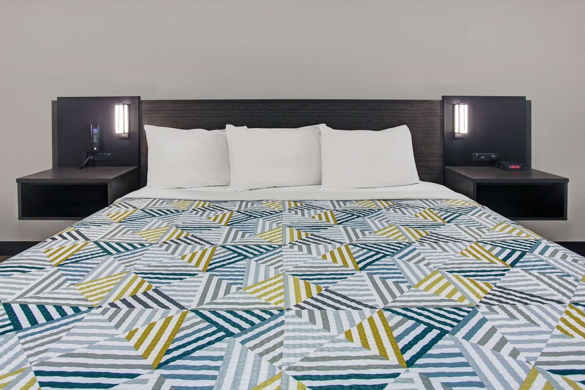 Une chambre d’hôtel avec un lit, des oreillers blancs, une couvre-lit à motifs géométriques et deux tables de chevet dotées d’un éclairage intégré.