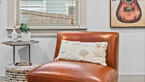 Un fauteuil en cuir marron avec un coussin blanc, une petite table ronde ornée d’une plante et d’une bouteille, et un tableau représentant une guitare, encadré, accroché au mur.
