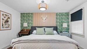 Une chambre à coucher comprenant un lit, deux tables de chevet, une décoration murale représentant une tête de cerf, une estampe botanique encadrée et une fenêtre avec des stores.
