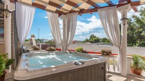 Un jacuzzi bajo una pérgola con cortinas blancas, rodeado de plantas en macetas y un muro de piedra.