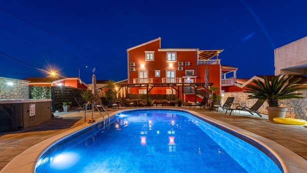 A swimming pool with a hot tub and lounge chairs, surrounded by a stone wall and a multi-story building with balconies.