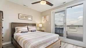 Une chambre avec un lit en bois, un ventilateur de plafond et une grande porte-fenêtre coulissante donnant sur un balcon.