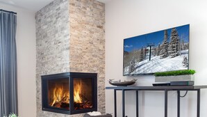 A modern living room with a stone fireplace, a glass coffee table with a magazine, a side table with a plant, and a painting of a snowy mountain scene. - Ski, Hike, Bike out the door. Hot Tub, Hot Pools, Gym, Pickleball,Tennis Courts (Park City)