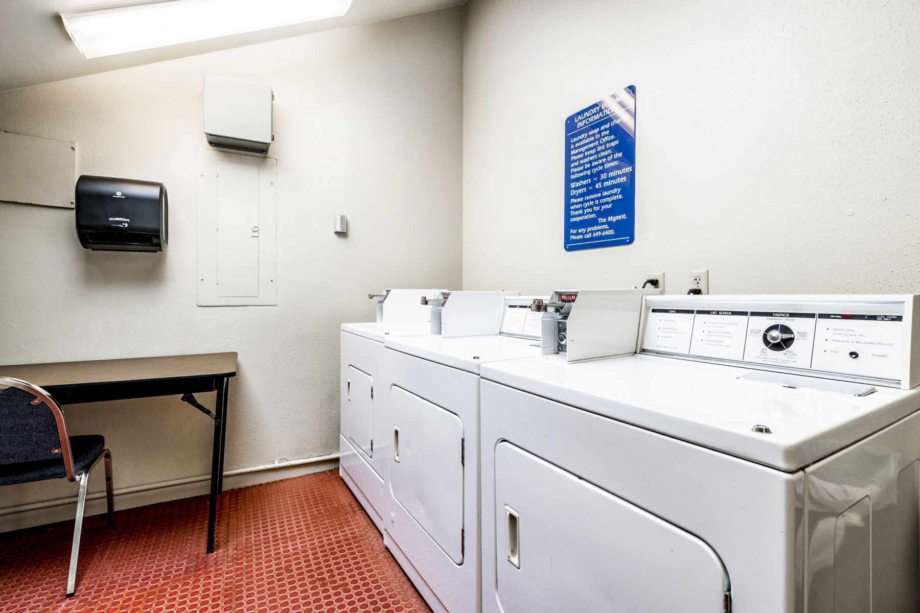 A laundry room with a washing machine, dryer, and a table with a chair. A sign provides laundry information.