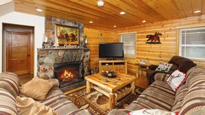 L’intérieur d’un chalet confortable, avec des murs en rondins de bois, une cheminée en pierre, un téléviseur et un coin salon accueillant.
