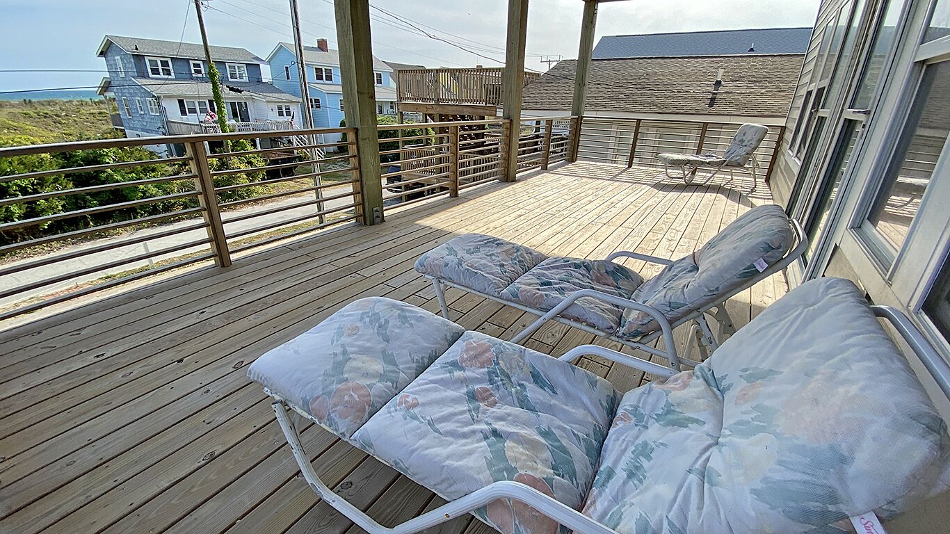 Une terrasse en bois avec deux chaises longues et une table, d’où l’on a vue sur une plage et des maisons.
