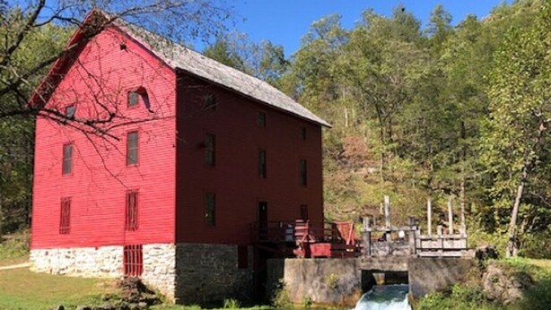 A red building with a stone foundation, a small bridge over a flowing river, and lush green trees in the background.