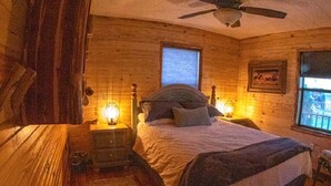 A cozy bedroom with a bed, wooden walls, a ceiling fan, and two windows.