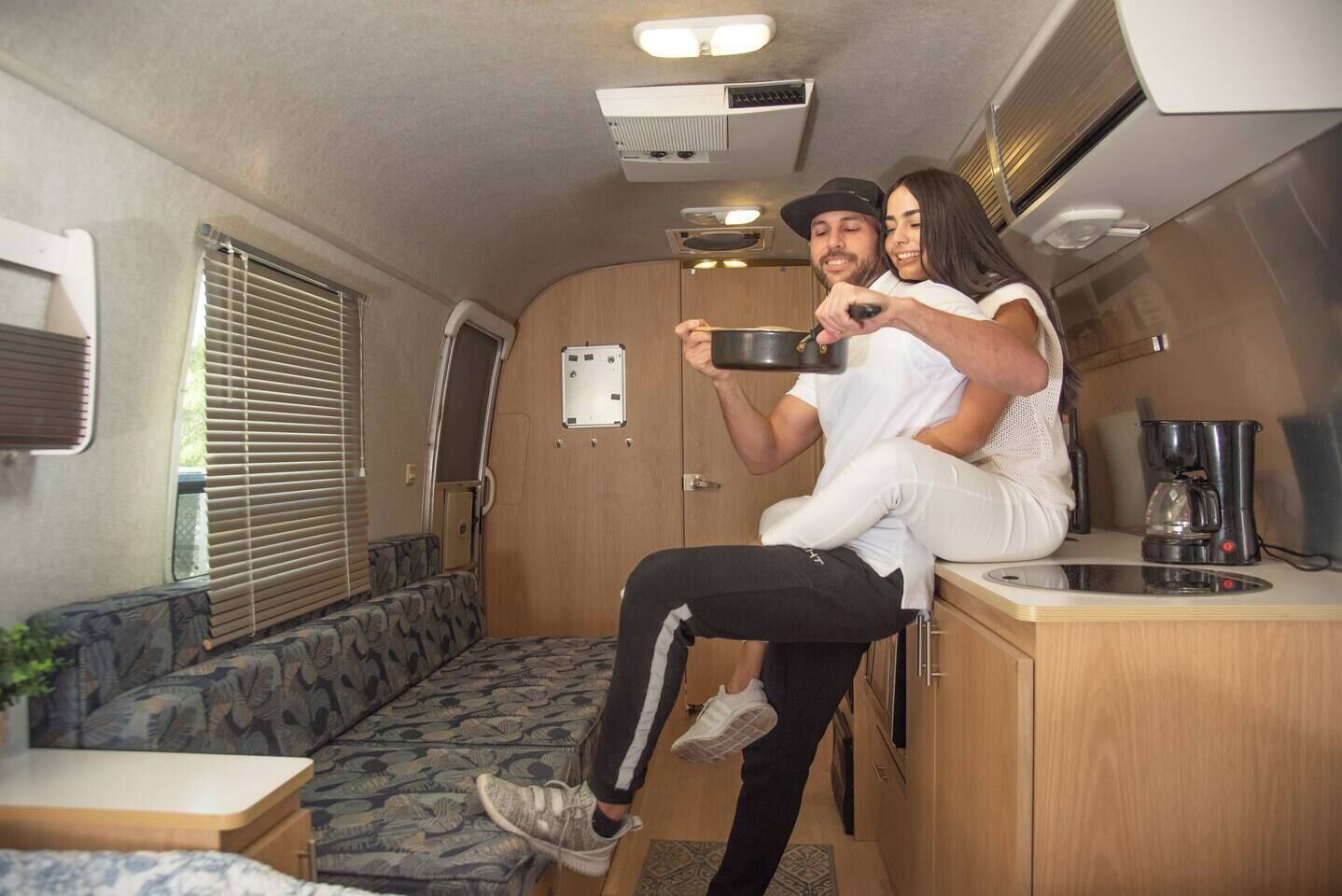 A compact RV interior with a kitchenette, seating area, and a window with blinds.