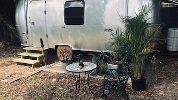 A silver trailer with a small table and chairs, surrounded by potted plants and a wooden bench.