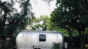 En sølvfarvet campingvogn parkeret i en baghave med en flisebelagt terrasse, omgivet af træer og plantekasser.