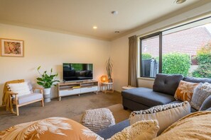 Ein Wohnzimmer mit einer Couch, einem Fernseher, einer Pflanze und einem Fenster mit Blick auf ein Haus.