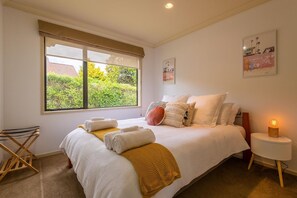 A neatly made bed with pillows and a blanket, a bedside lamp, and a window with a view of greenery. - Iles Inn - Spa Pool, Pets OK, Walk to Forest (Rotorua)