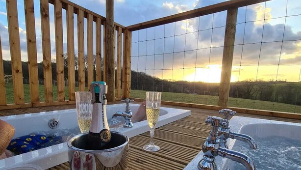 A hot tub with champagne and glasses, surrounded by wooden deck and a scenic view. - Luxury Safari Lodge-Sleeps 4/6-Deer Park-Dogs Ok (Crediton)